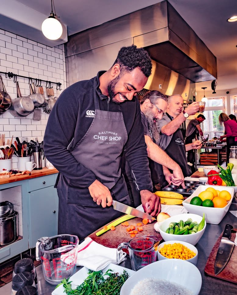 Parrish laughing while chopping vegetables during a hands-on cooking class at Baltimore Chef Shop, a teaching kitchen, surrounded by fresh produce